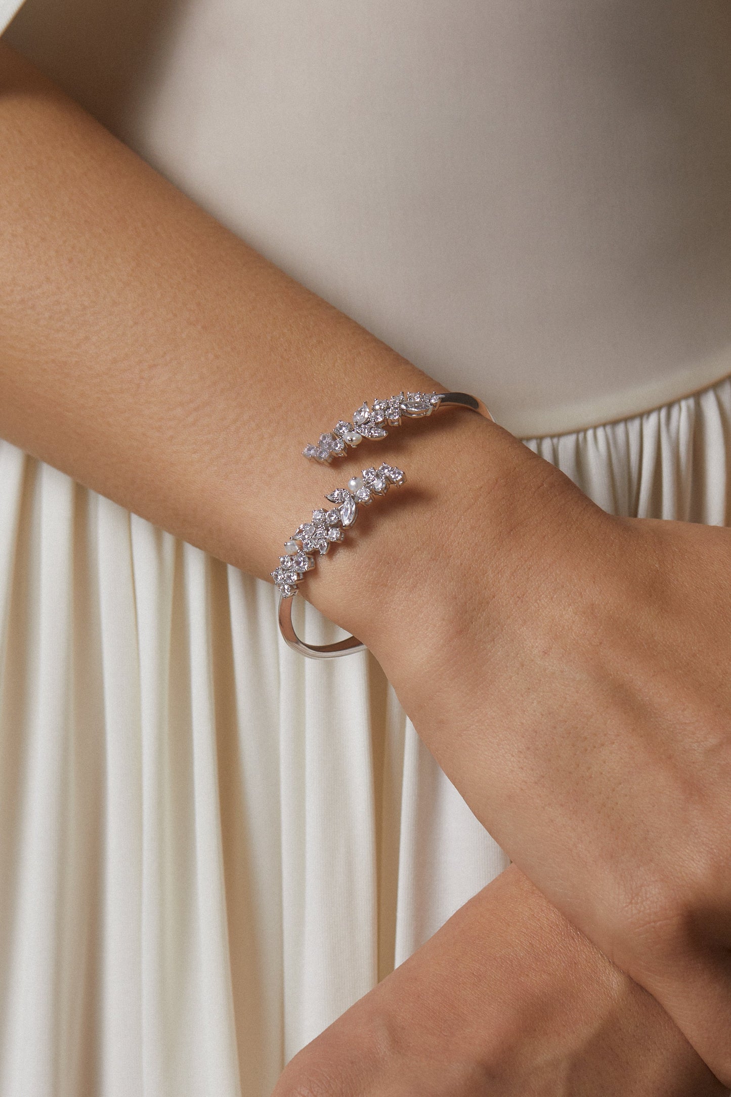 MODEL WEARING RAVISH LAUREL BYPASS BRACELET. IT IS A CLOSE UP IMAGE OF HER WRISTS WITH A WHITE DRESS IN THE BACKGROUND. 