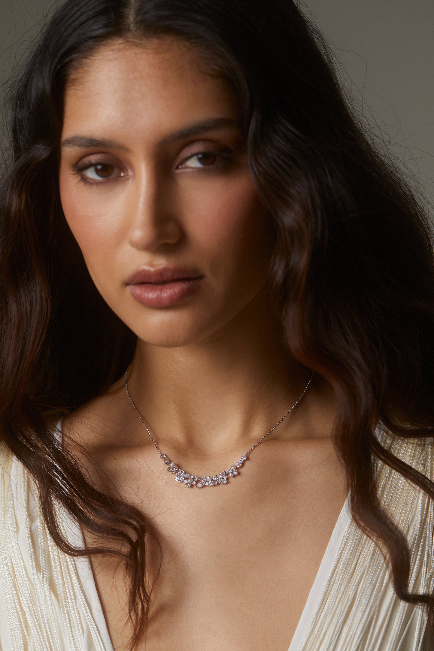 MODEL WEARING RAVISH LAUREL FRONTAL NECKLACE. SHE IS LOOKING DIRECTLY INTO THE CAMERA AND WEARING A WHITE PLEATED VNECK TOP AND HER LONG DARK BROWN HAIR BLOWING IN THE WIND. 