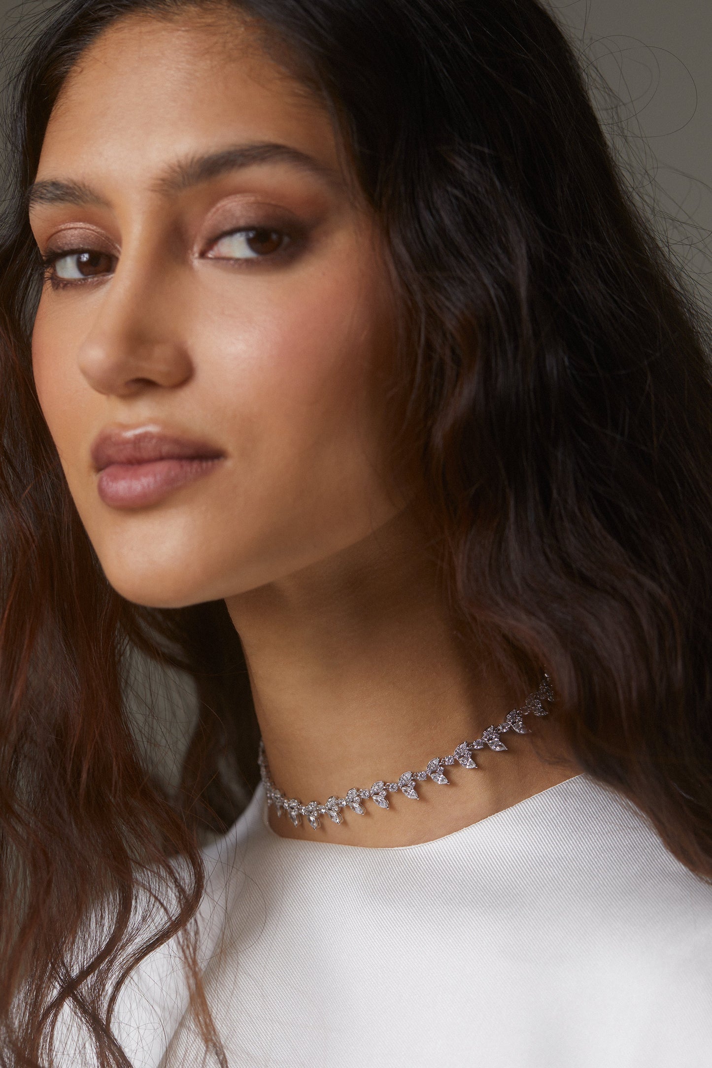 MODEL WEARING RAVISH HALF VINE NECKLACE AS A CHOKER. SHE IS TURNED TOWARDS THE CAMERA WITH HER DARK BROWN HAIR FLOWING AROUND HER FACE. SHE IS WEARING A HIGH NECKLINE WHITE TOP. 