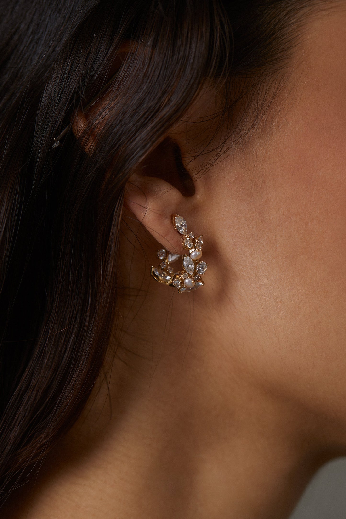 RAVISH MINI C HOOP EARRINGS ON MODEL. IT IS A CLOSE UP IMAGE OF HER EAR WITH HER FACE CROPPED OUT. HER DARK BROWN HAIR IS FALLING OVER HER EARS BUT THE SIDE OF THE EARRINGS ARE VISIBLE. 