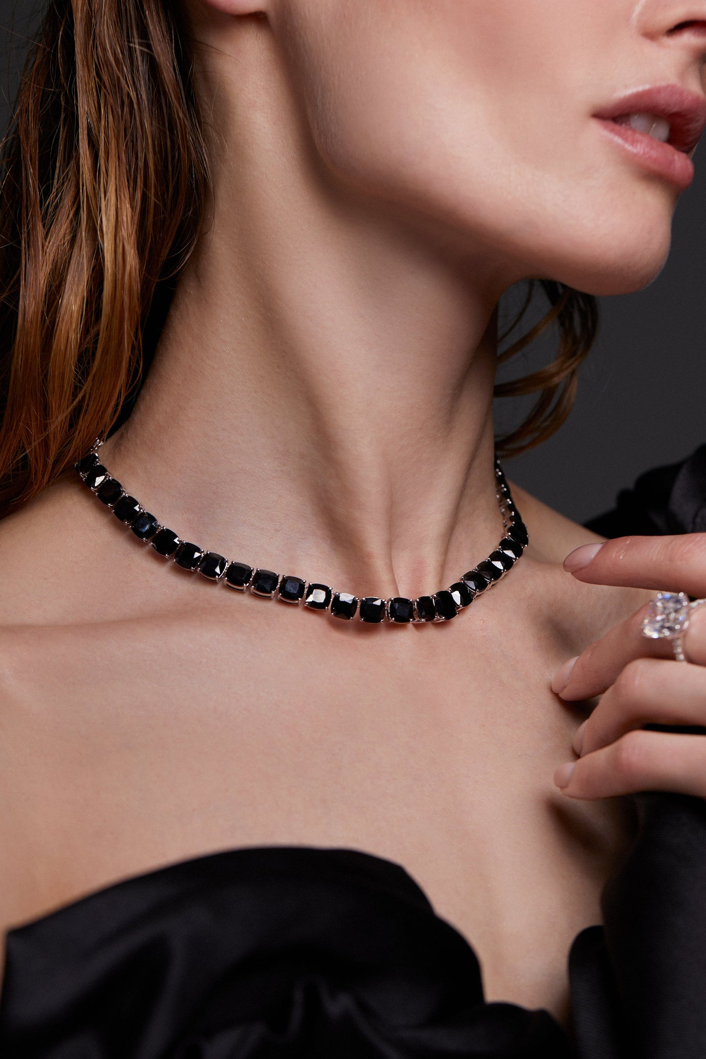 MODEL WEARING STARLET BLACK CUSHION COLLAR NECKLACE. IT IS A CLOSE UP IMAGE OF THE MODELS NECK. HER HAIR IS BEHIND HER SHOULDERS AND HER FINGERTIPS ARE REACHING FOR THE NECKLACE. 