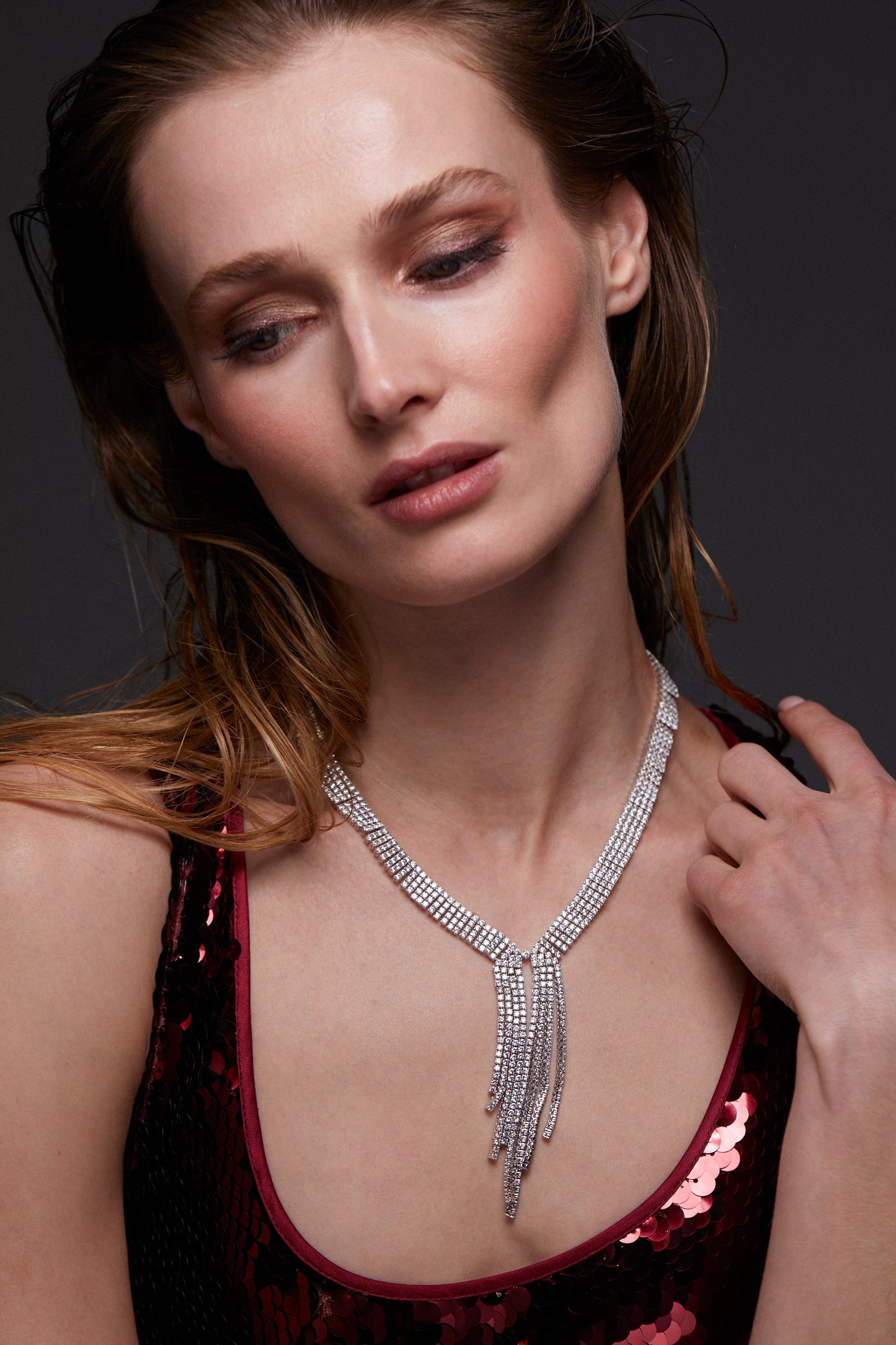 MODEL WEARING STARLET DRAMA Y NECKLACE. SHE IS LOOKING DOWN AND HER HAIR IS TOUSLED BEHIND HER SHOULDER. SHE IS WEARING A RED SEQUIN DRESS. 