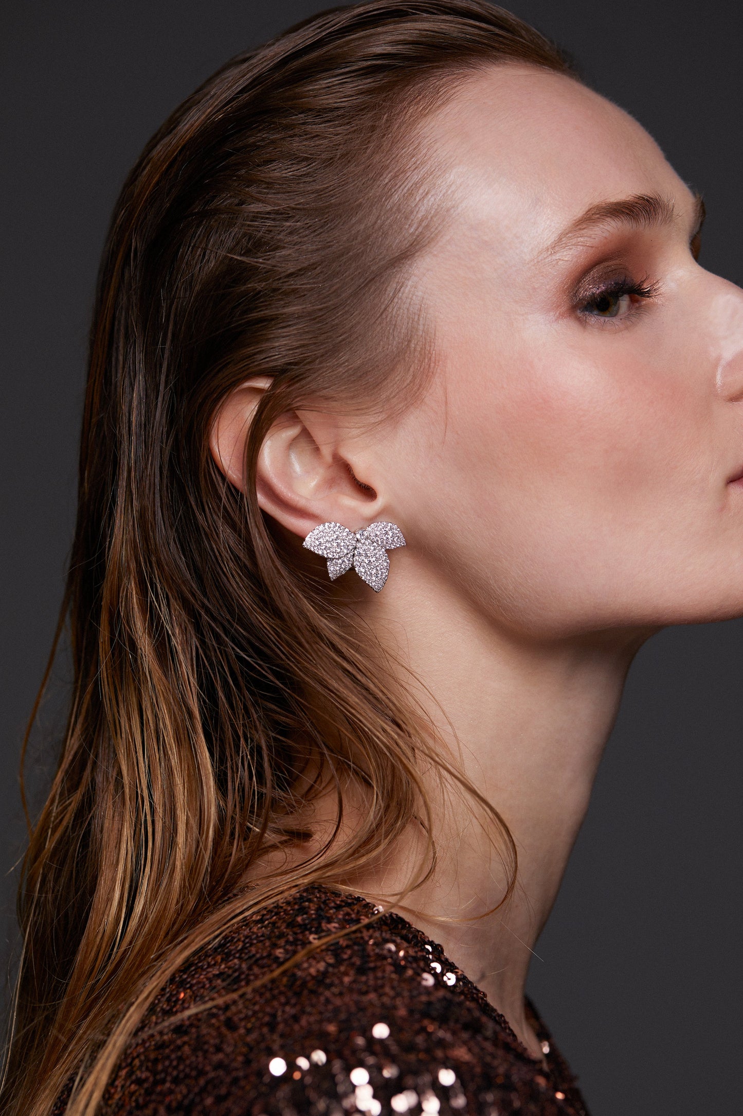 MODEL WEARING STARLET LEAF STUD EARRINGS. SHE IS FACING TO THE SIDE WITH HER HAIR SLICKED BACK AND TUCKED BEHIND HER EARS. SHE IS WEARING A SEQUIN ROSE GOLD METALLIC DRESS. 