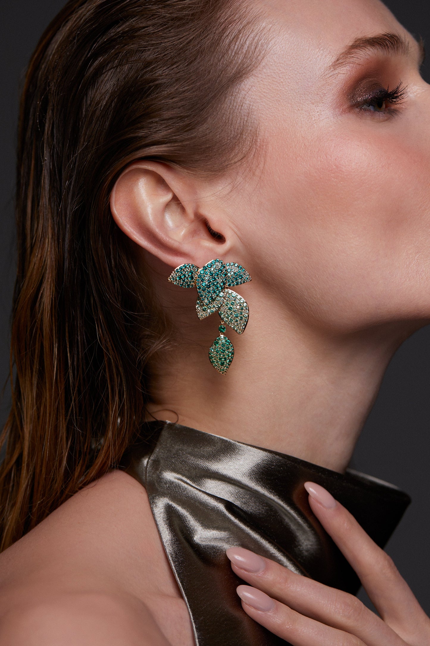 MODEL WEARING STARLET GREEN LEAF LINEAR EARRINGS. IT IS A CLOSE UP IMAGE WITH HER FACING TO THE RIGHT. HER HAIR IS TUCKED BEHIND HER EAR AND HER HAND IS REACHING UP FOR HER NECK. SHE IS WEARING A METALLIC GRAY HIGH NECK SLEEVELESS TOP. 