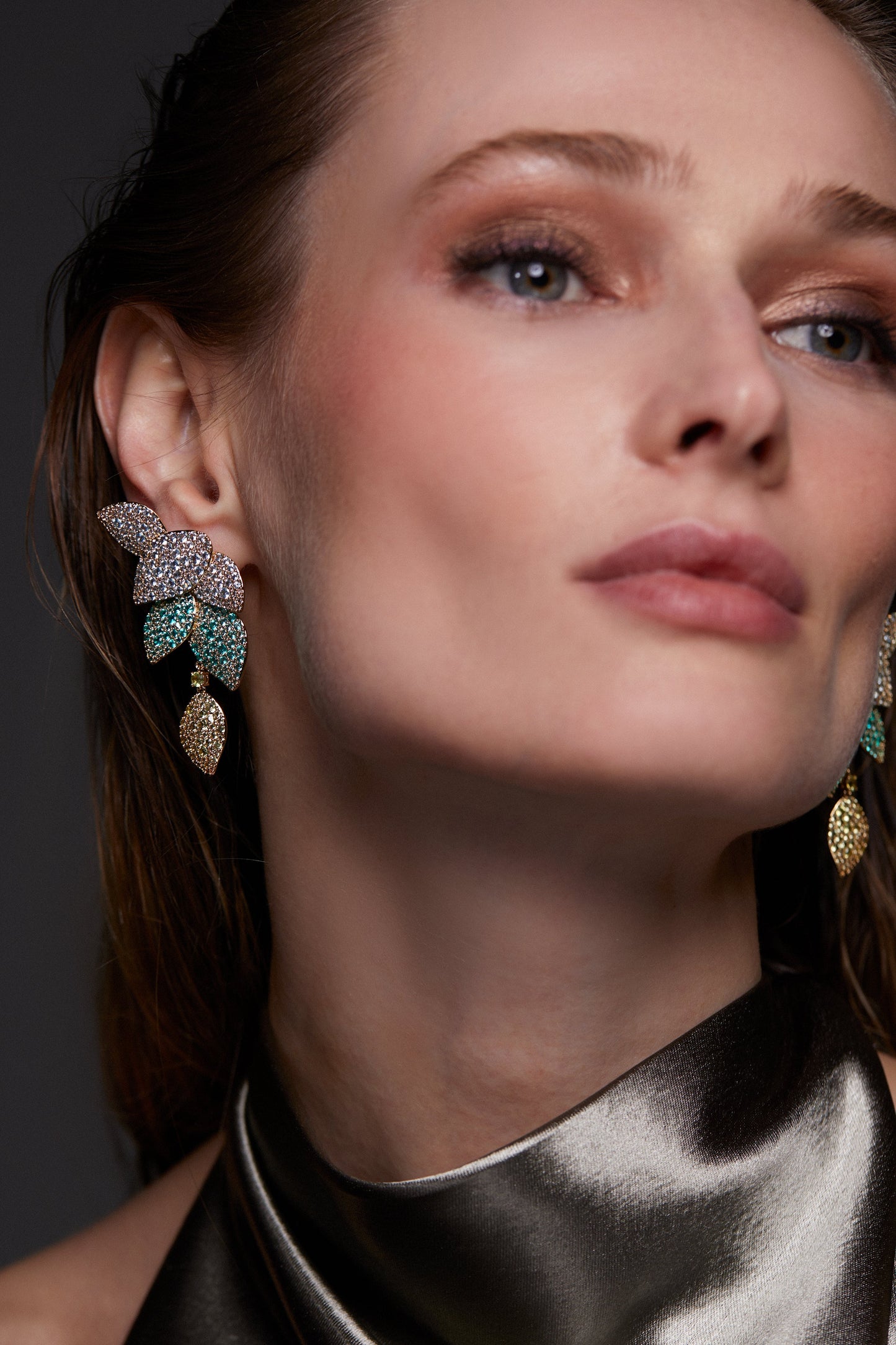 MODEL WEARING STARLET MULTI COLOR LEAF LINEAR EARRINGS. IT IS A CLOSE UP IMAGE WITH HER LOOKING TO THE RIGHT. SHE IS WEARING A METALLIC GRAY HIGH NECK SLEEVELESS TOP. 