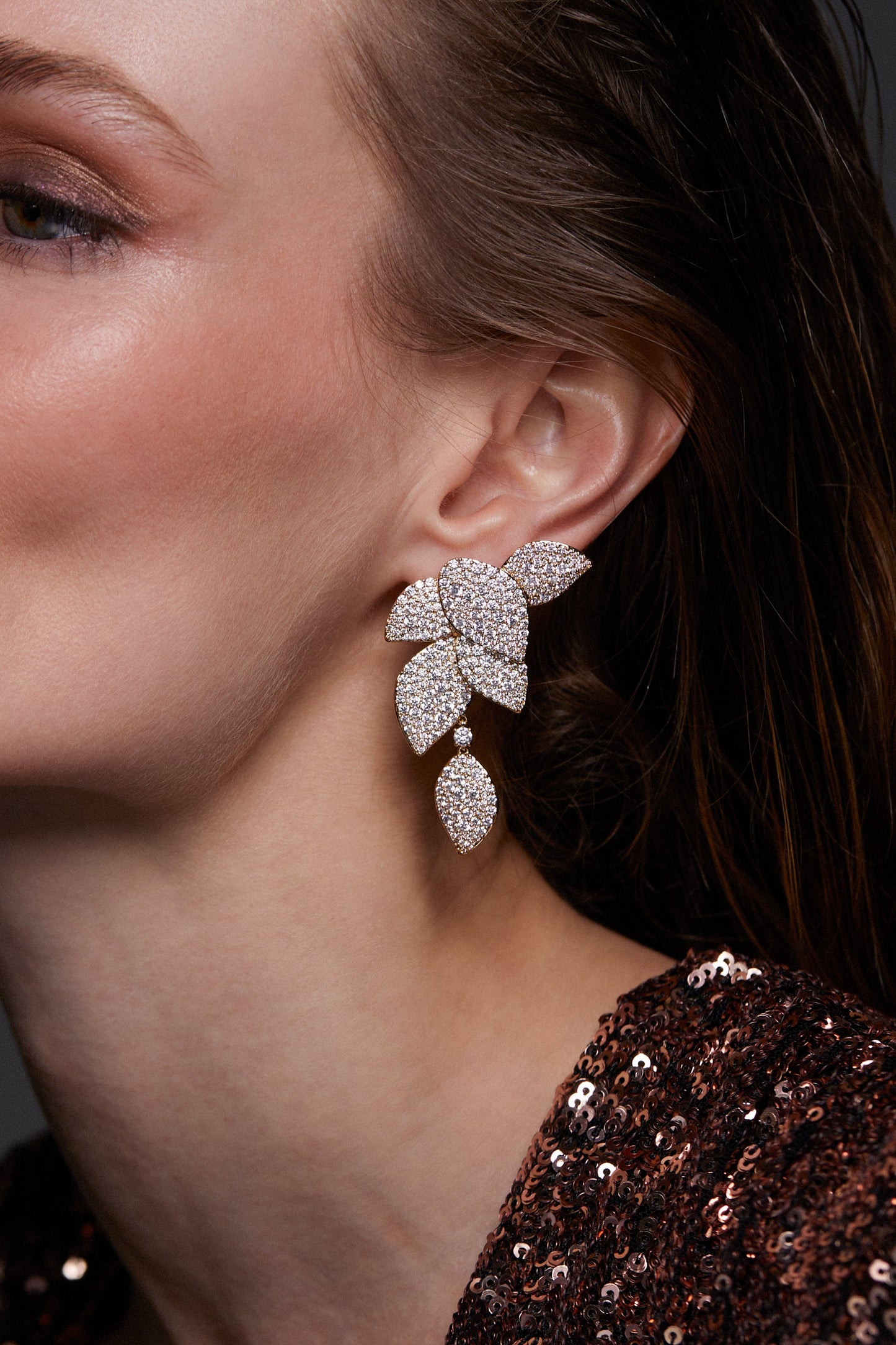 STARLET LEAF LINEAR EARRINGS ON MODEL. IT IS A CLOSE UP IMAGE OF HER EAR. HER HAIR IS TUCKED BEHIND HER EAR AND SHE IS WEARING A ROSE GOLD SEQUIN TOP. 