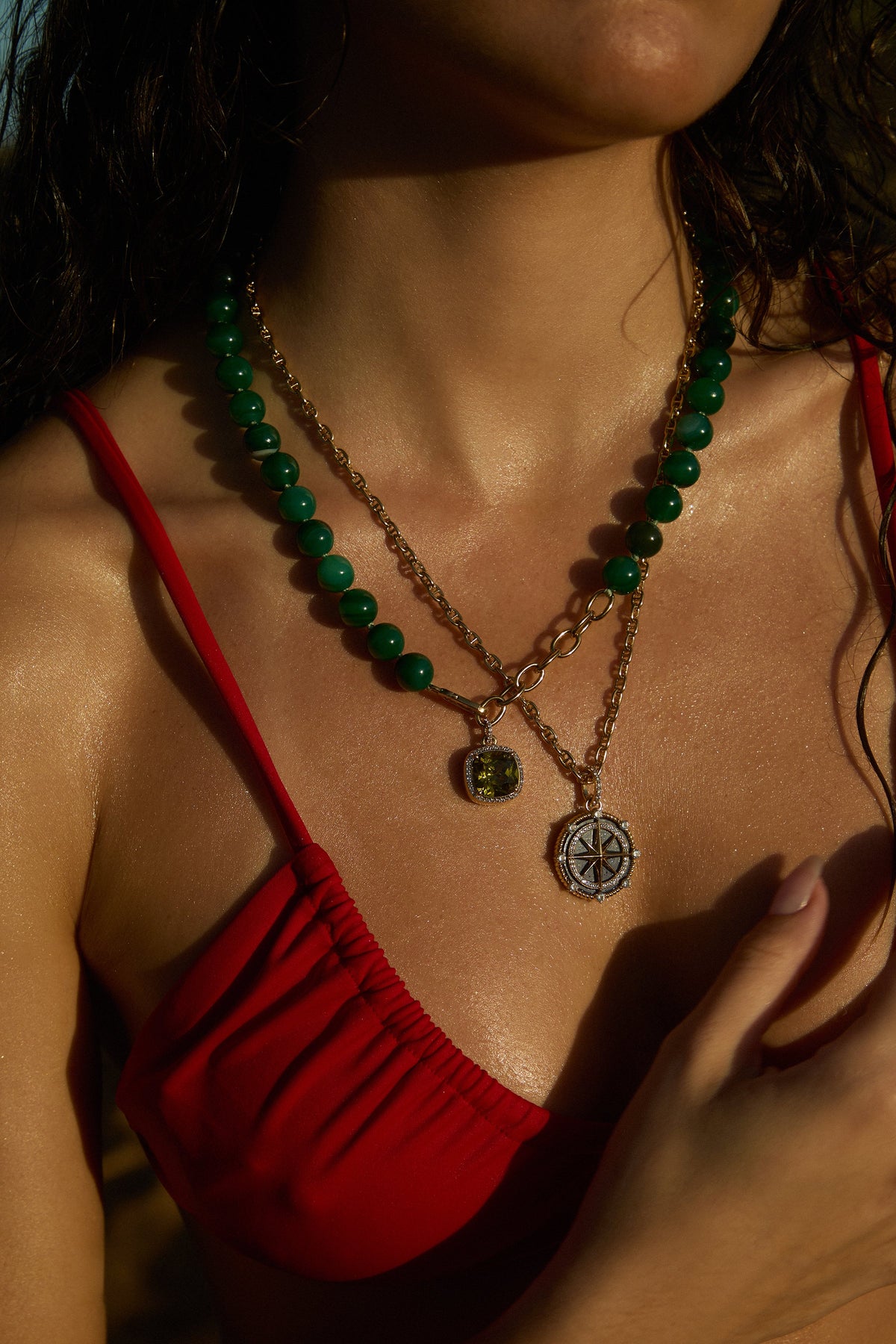 CLOSE UP OF A MODEL’S CHEST AS SHE WEARS A RED BATHING SUIT, THE BEADS OF PARADISE GREEN ADJUSTABLE Y NECKLACE, AND NAUTICAL PENDANT NECKLACE