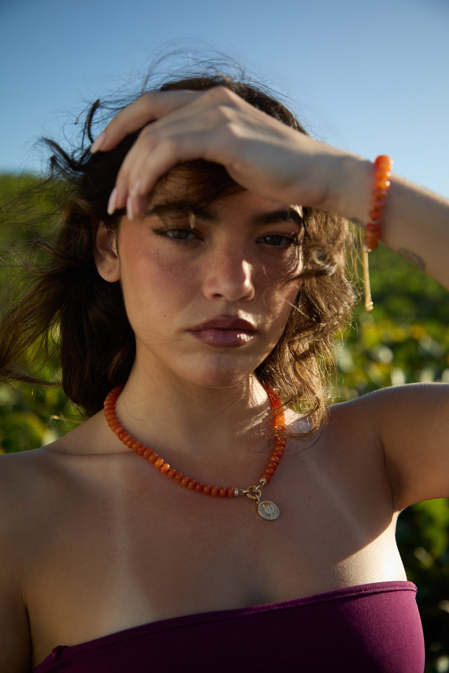MODEL OUTSIDE WITH HER HAIR BLOWING AND HAND LIFTED TO HER FOREHEAD, WEARING THE BEADS OF PARADISE SUN PENDANT NECKLACE AND MATCHING BOLO BRACELET 