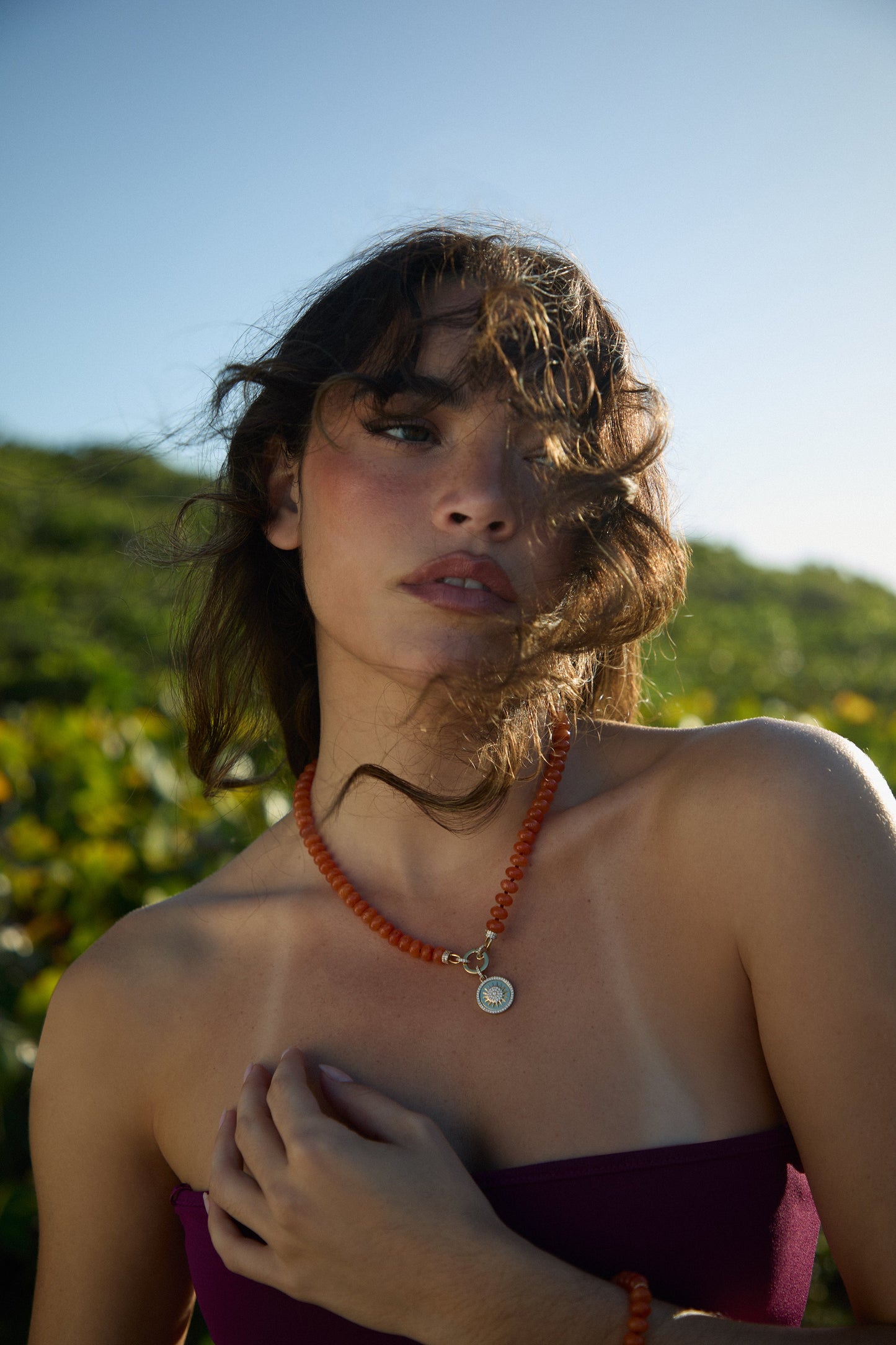 MODEL OUTSIDE WITH HER HAIR BLOWING IN FRONT OF HER FACE, WEARING A PURPLE BANDEAU TOP, THE BEADS OF PARADISE SUN PENDANT NECKLACE, AND MATCHING BOLO BRACELET 