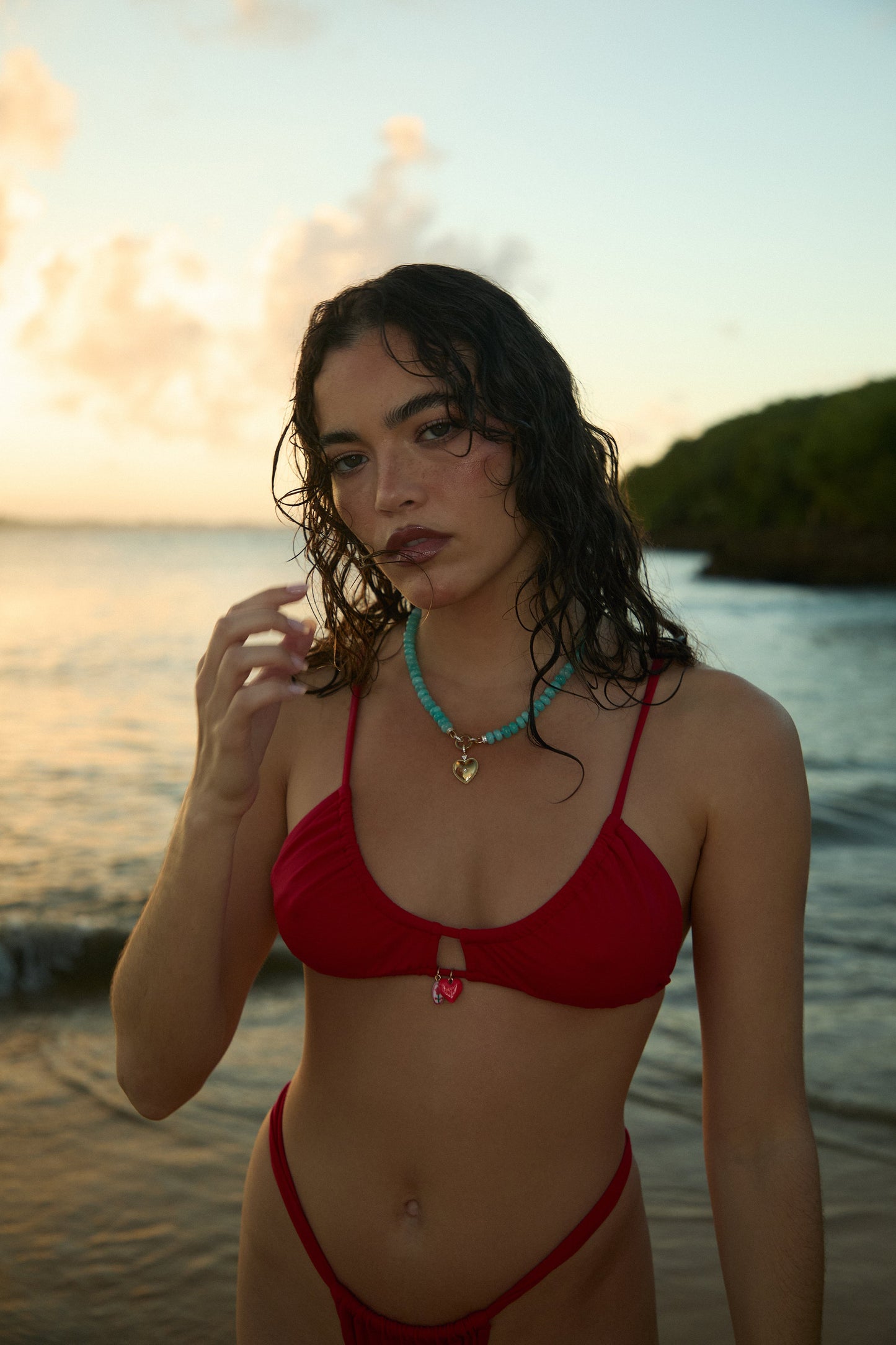 MODEL IN A RED BIKINI STANDING IN FRONT OF THE OCEAN, LIFTING HER HAND UP AND WEARING THE BEADS OF PARADISE HEART PENDANT NECKLACE