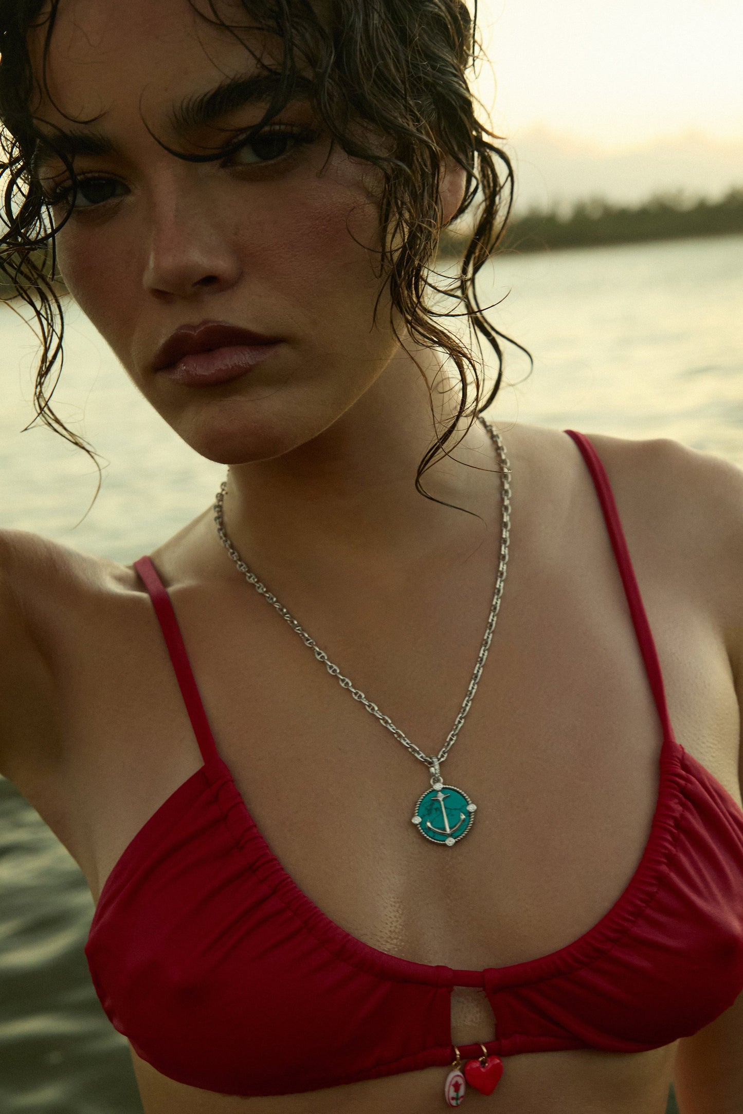 CLOSE UP OF A MODEL IN A RED BATHING SUIT IN THE OCEAN, HOLDING HER HAIR UP AS SHE WEARS THE GLOBE TROTTER TURQUOISE ANCHOR PENDANT NECKLACE