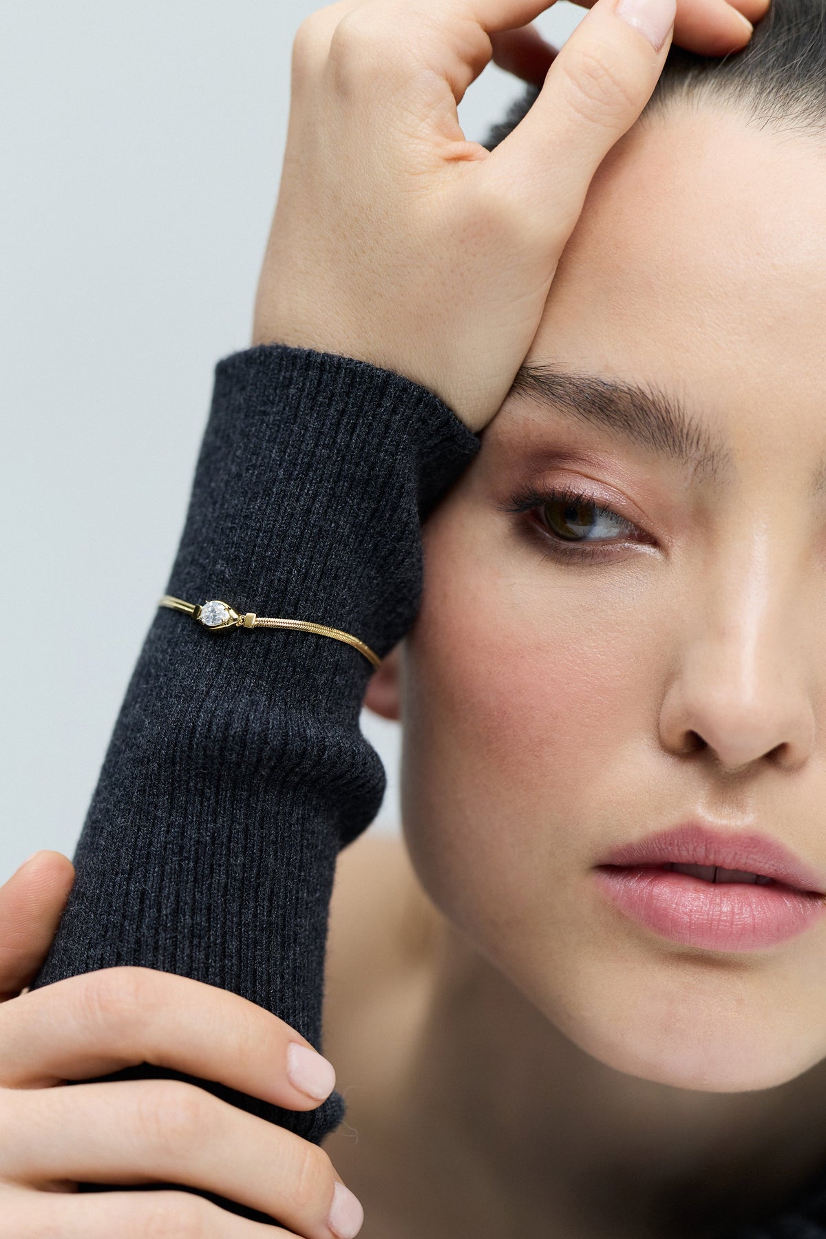 CLOSE UP OF A MODEL’S WRIST LEANING ON HER HEAD, AS SHE WEARS THE DELANCEY PEAR LINE BRACELET LAYERED OVER A GRAY SWEATER