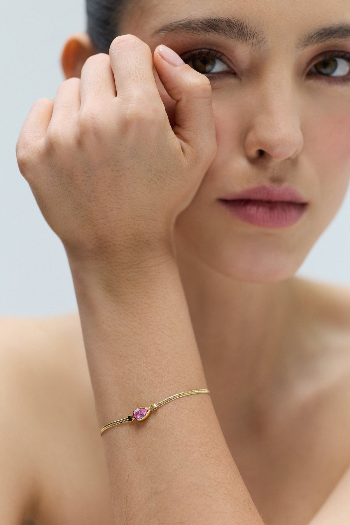 CLOSE UP OF A MODEL LOOKING AT THE CAMERA WITH HER WRIST LIFTED UP TO HER FACE, WEARING THE DELANCEY PINK PEAR LINE BRACELET