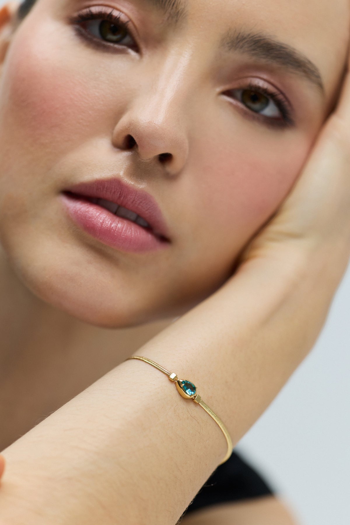 CLOSE UP OF A MODEL’S FACE LOOKING AT THE CAMERA, WITH HER HEAD LEANING ON HER WRIST AS SHE WEARS THE DELANCEY AQUA PEAR LINE BRACELET