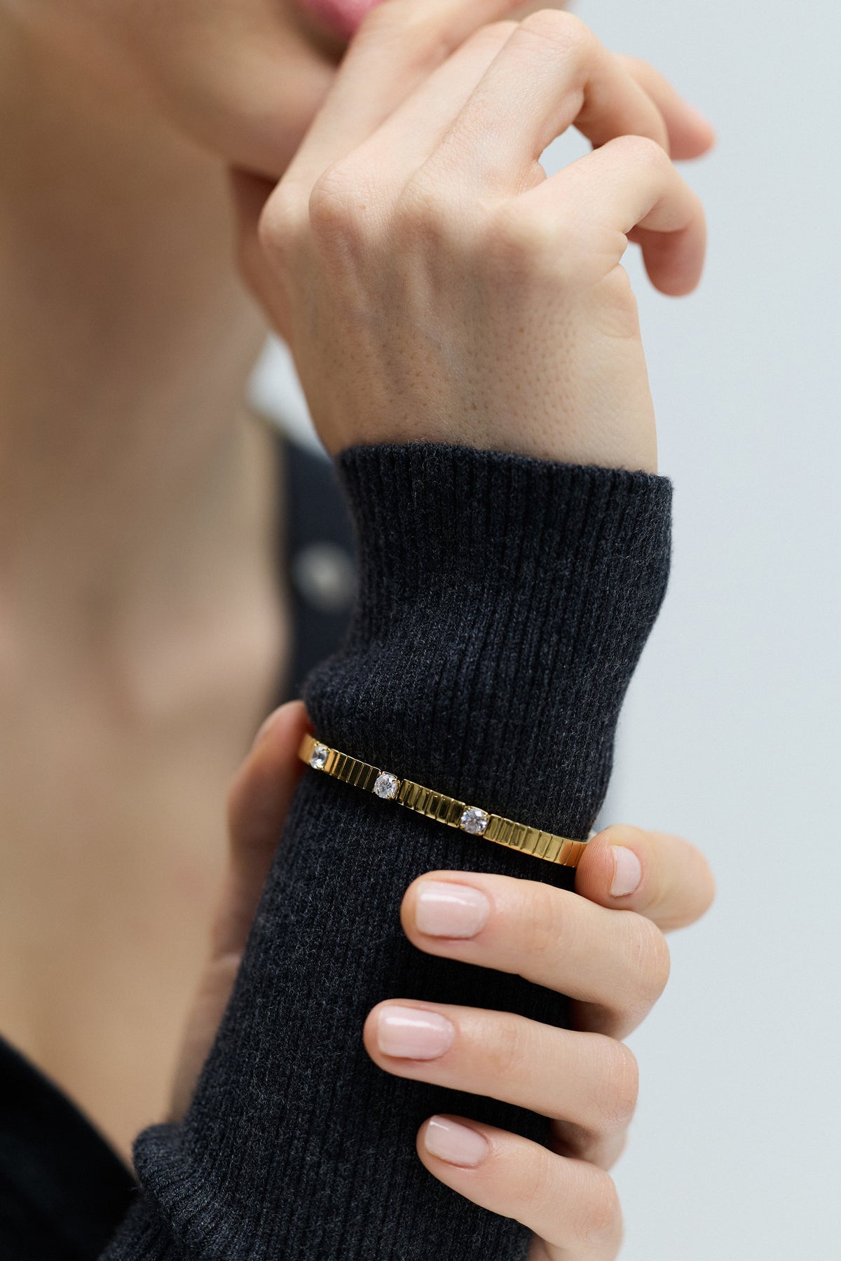 CLOSE UP OF A MODEL LIFTING HER WRIST AND HOLDING IT WITH THE OTHER HAND, AS SHE WEARS THE DELANCEY BOTTOM HINGE BRACELET LAYERED OVER A GRAY SWEATER
