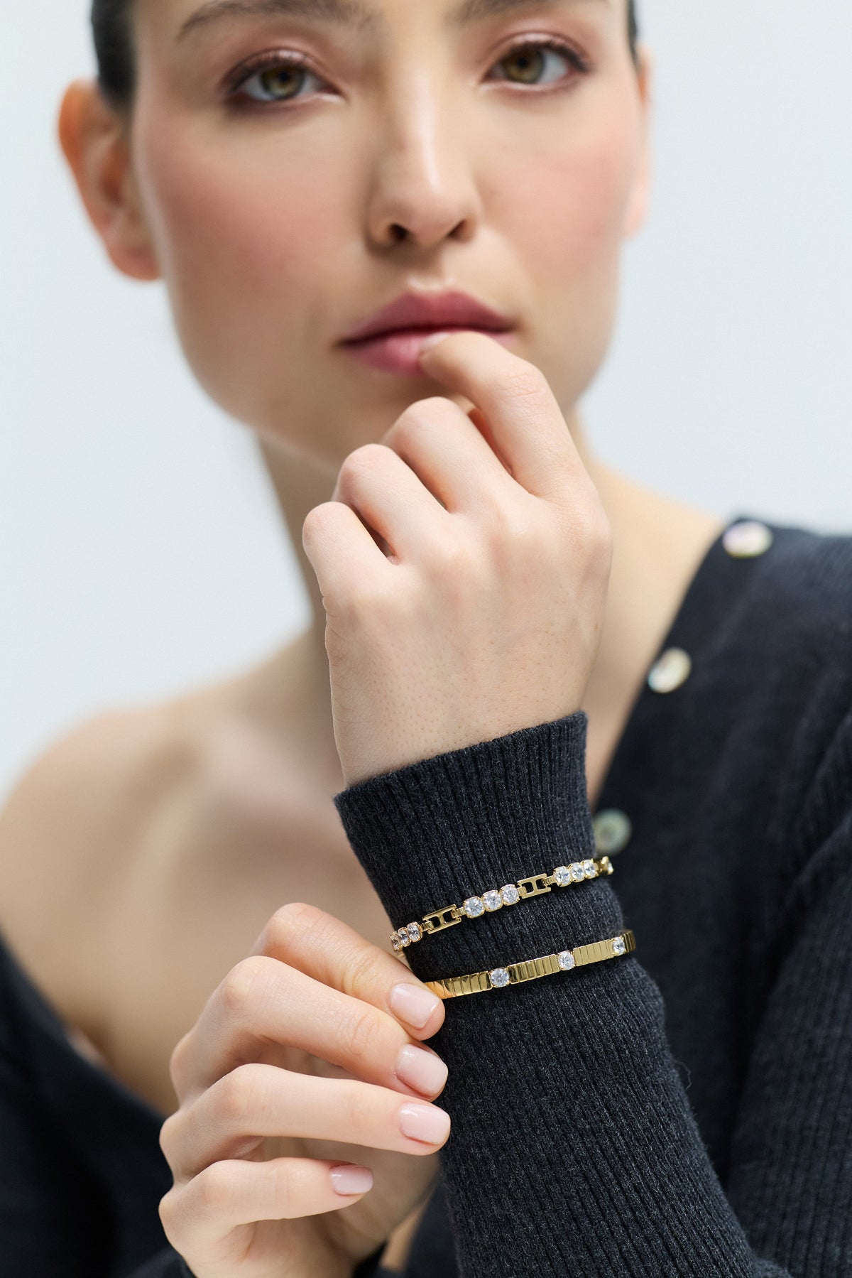 A MODEL LIFTING HER WRIST IN FRONT OF HER, AS SHE WEARS THE DELANCEY BOTTOM HINGE BRACELET AND THE DELANCEY LINE BRACELET LAYERED OVER A GRAY SWEATER