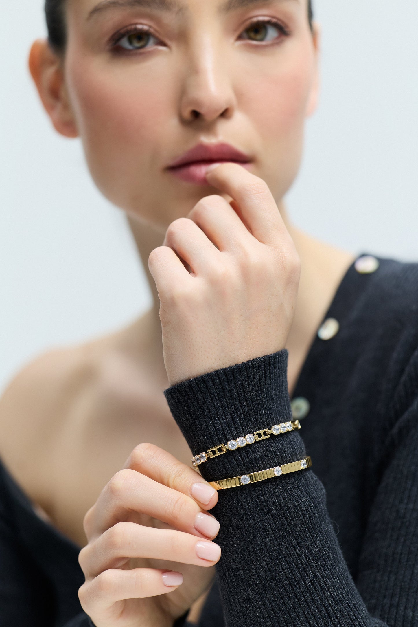 A MODEL LIFTING HER WRIST IN FRONT OF HER, AS SHE WEARS THE DELANCEY BOTTOM HINGE BRACELET AND THE DELANCEY LINE BRACELET LAYERED OVER A GRAY SWEATER