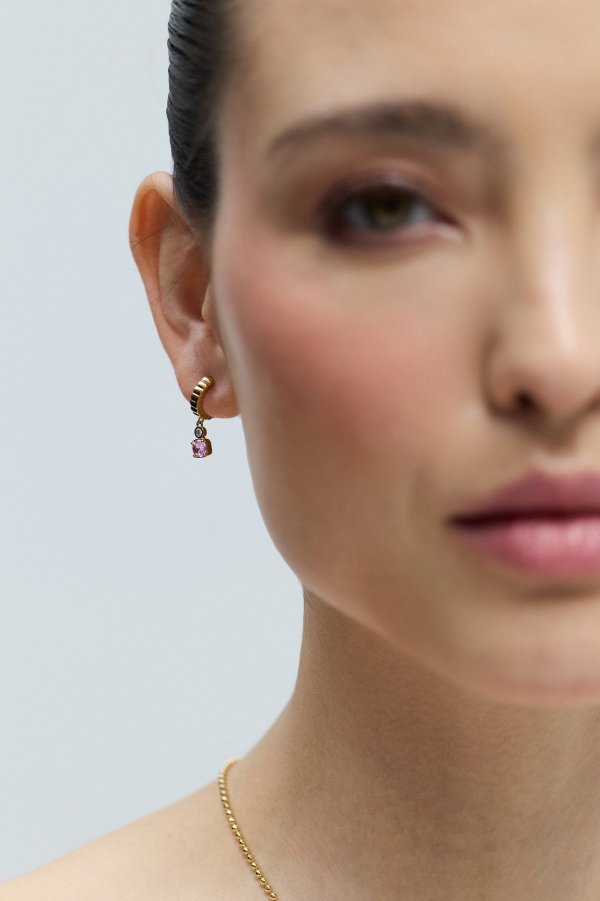 CLOSE UP OF HALF OF A MODEL’S FACE WITH ONLY HER EAR IN FOCUS, AS SHE WEARS THE DELANCEY PINK SMALL CHARM HOOP EARRINGS