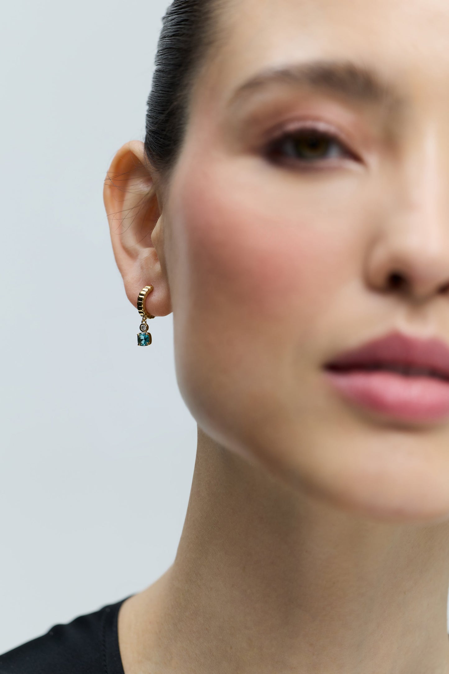 CLOSE-UP OF HALF OF A MODEL'S FACE WITH HER EAR IN FOCUS, WEARING THE DELANCEY AQUA SMALL CHARM HOOP EARRINGS.