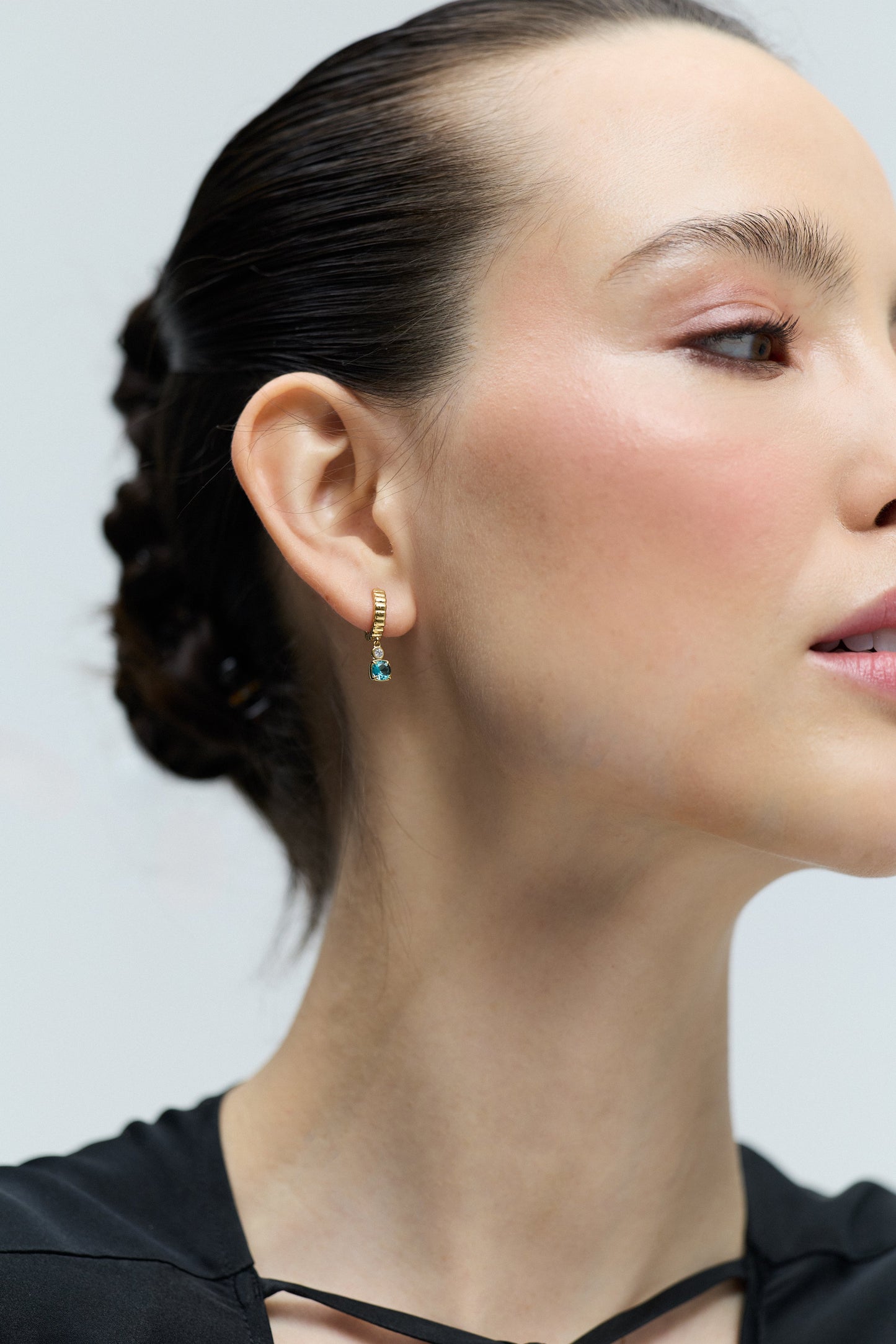 CLOSE UP OF A MODEL WITH AN UPDO HAIRSTYLE TURNED TO THE SIDE, WEARING THE DELANCEY AQUA SMALL CHARM HOOP EARRINGS