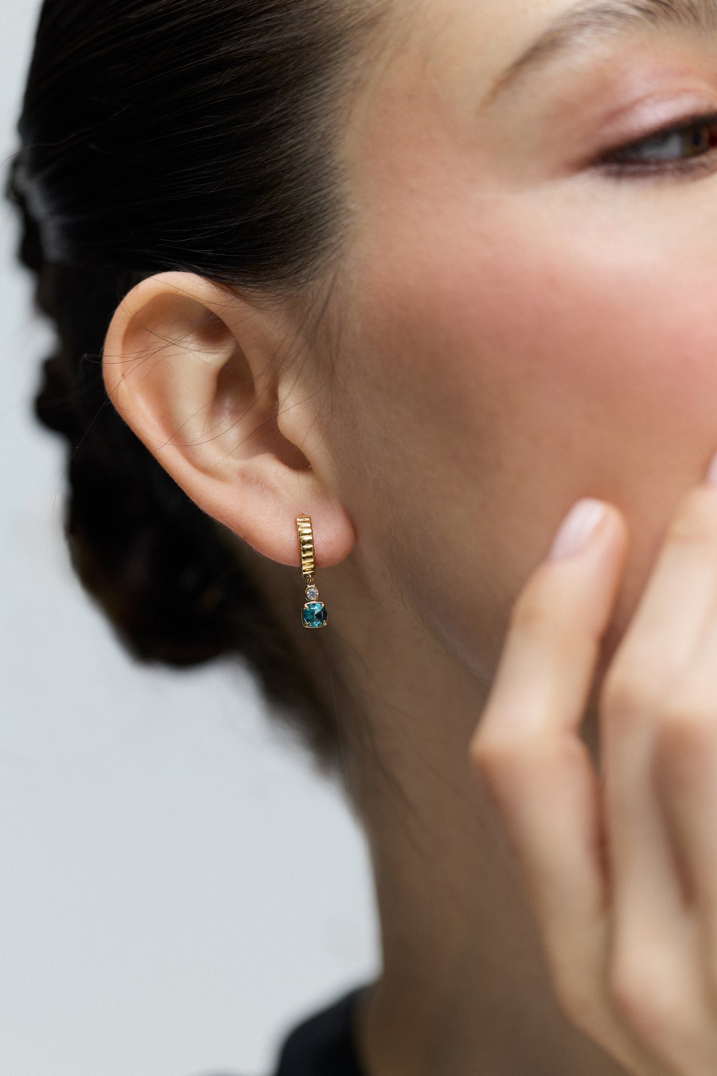 CLOSE UP OF A MODEL TURNED TO THE SIDE WITH HER EAR IN FOCUS, AS SHE WEARS THE DELANCEY AQUA SMALL CHARM HOOP EARRINGS