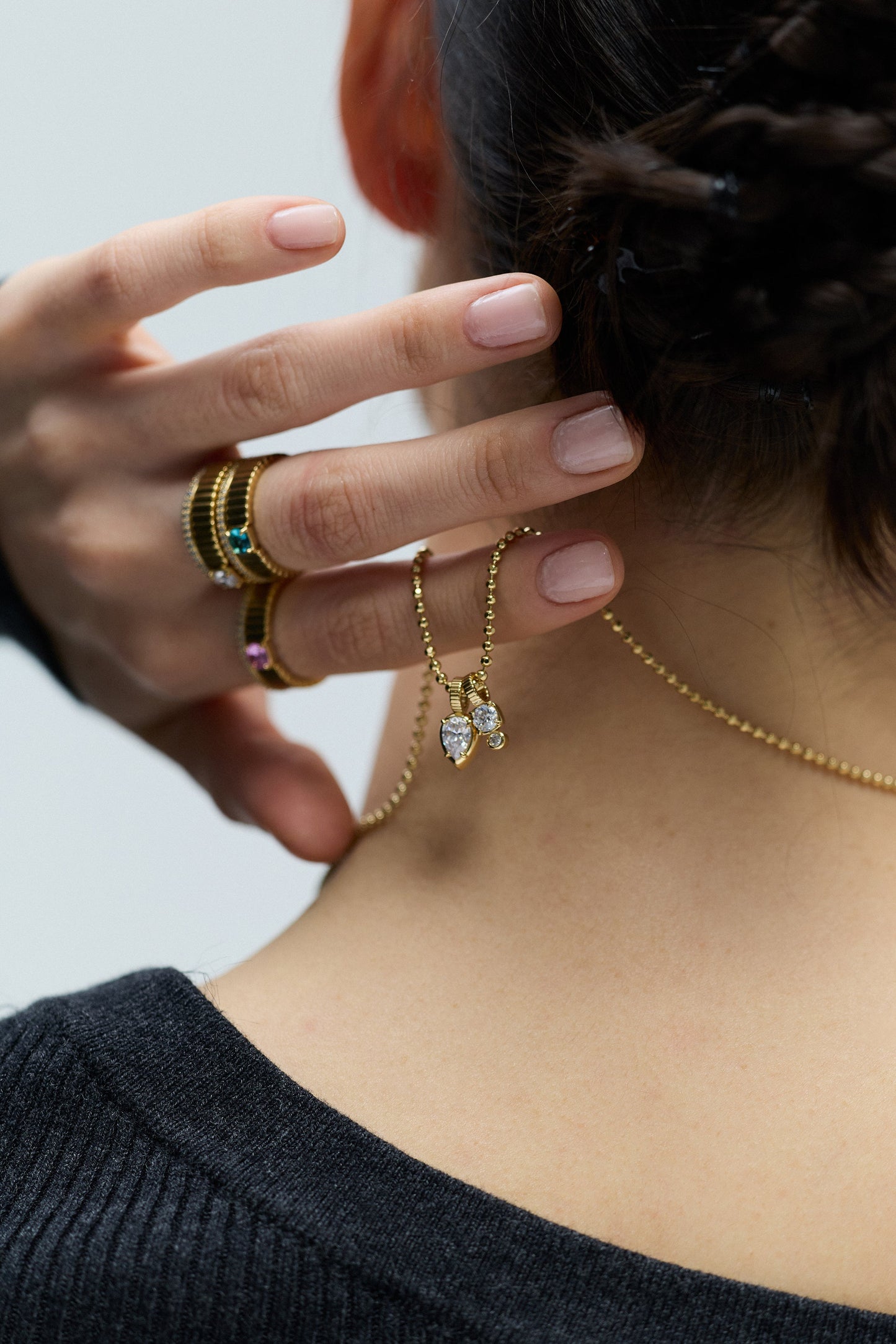 BACK VIEW OF A MODEL WITH A TWISTED BRAID UPDO, HOLDING THE DELANCEY CHARM PENDANT NECKLACE AT THE BACK OF HER NECK, WHILE WEARING DELANCEY SOLITAIRE RINGS ON HER FINGERS