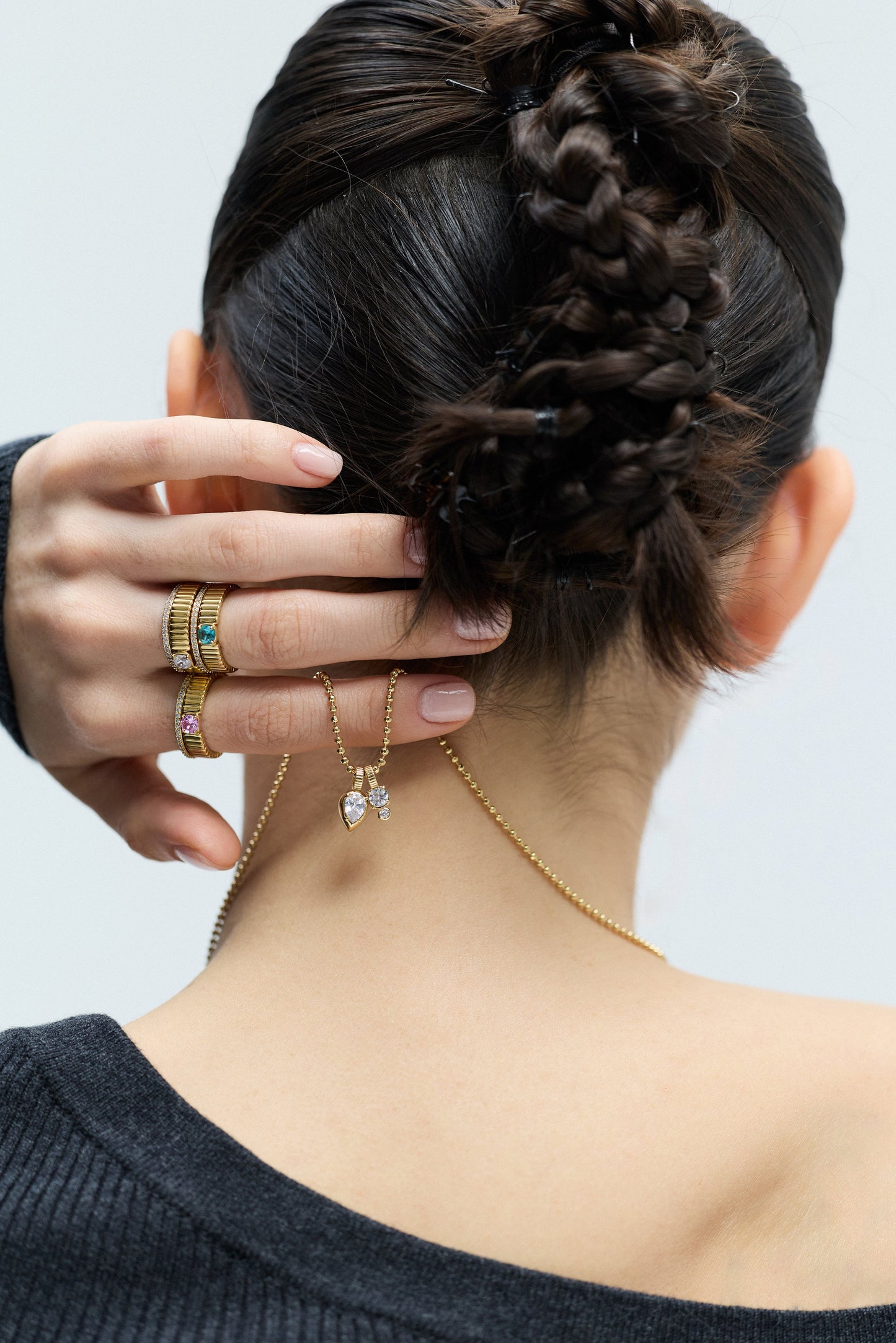 BACK VIEW OF A MODEL WITH A TWISTED BRAID UPDO, HOLDING THE DELANCEY CHARM PENDANT NECKLACE AT THE BACK OF HER NECK, WHILE WEARING DELANCEY SOLITAIRE RINGS ON HER FINGERS