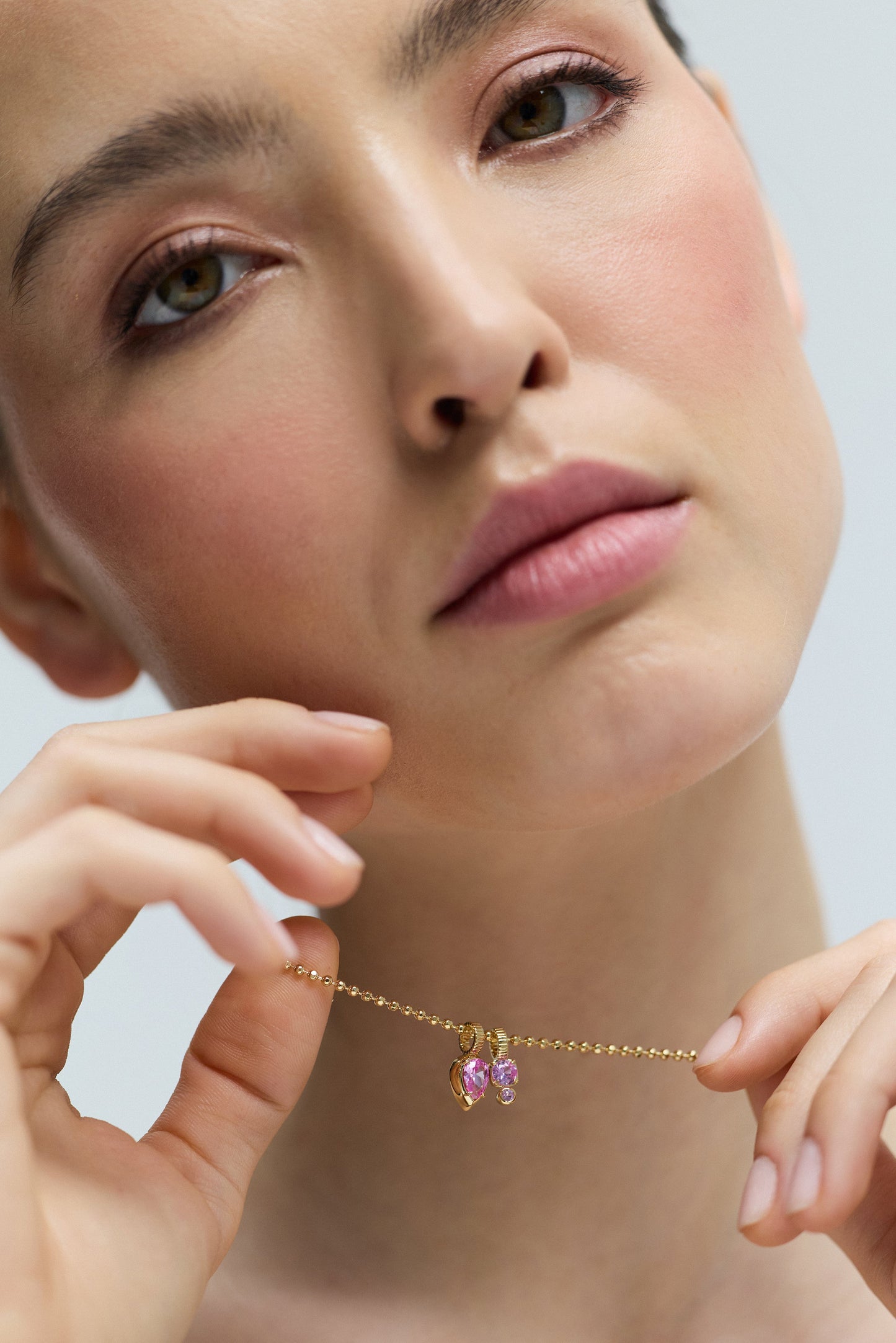 CLOSE UP OF A MODEL’S FACE, AS SHE WEARS THE DELANCEY PINK CHARM PENDANT NECKLACE AND LIFTS IT UP TO HER CHIN