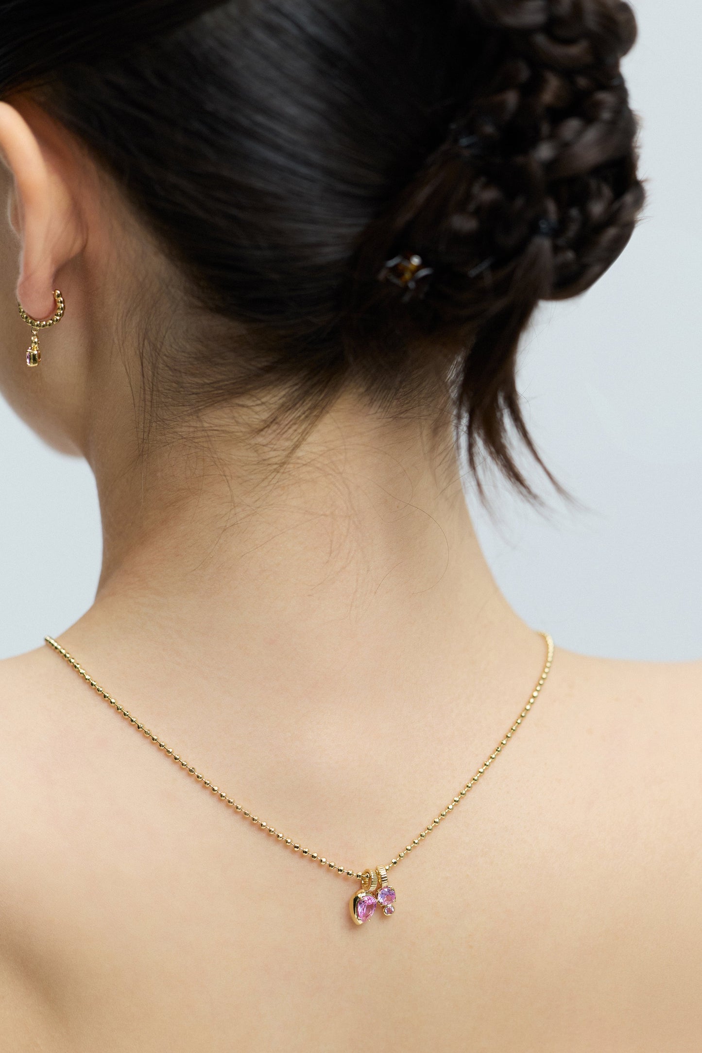 CLOSE UP OF THE BACK OF A MODEL’S HEAD WITH A TWISTED BRAID UPDO, WEARING THE DELANCEY PINK CHARM PENDANT NECKLACE STYLED BACKWARDS SO IT DRAPES DOWN HER BACK