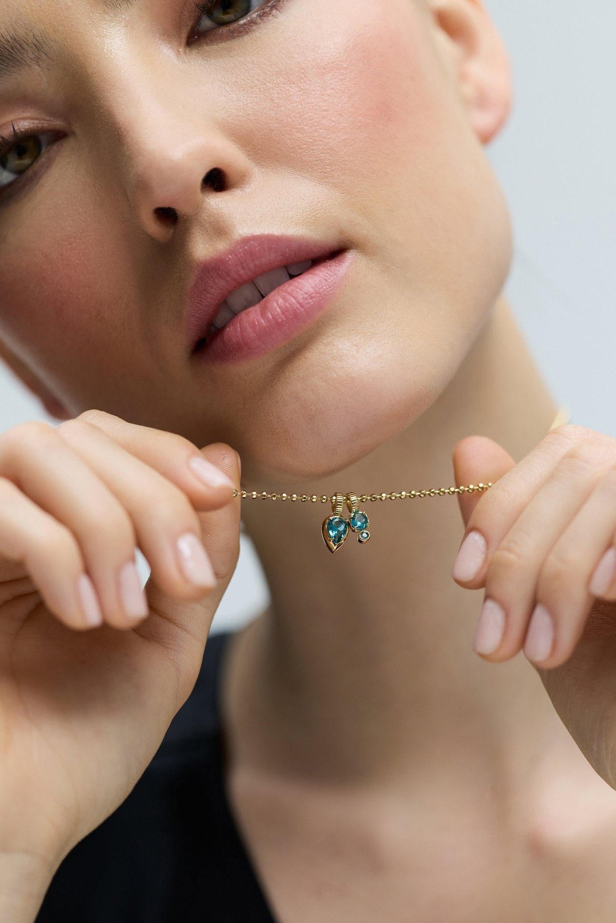 CLOSE UP OF A MODEL’S FACE AS SHE WEARS THE DELANCEY AQUA CHARM PENDANT NECKLACE AND LIFTS IT UP TO HER CHIN