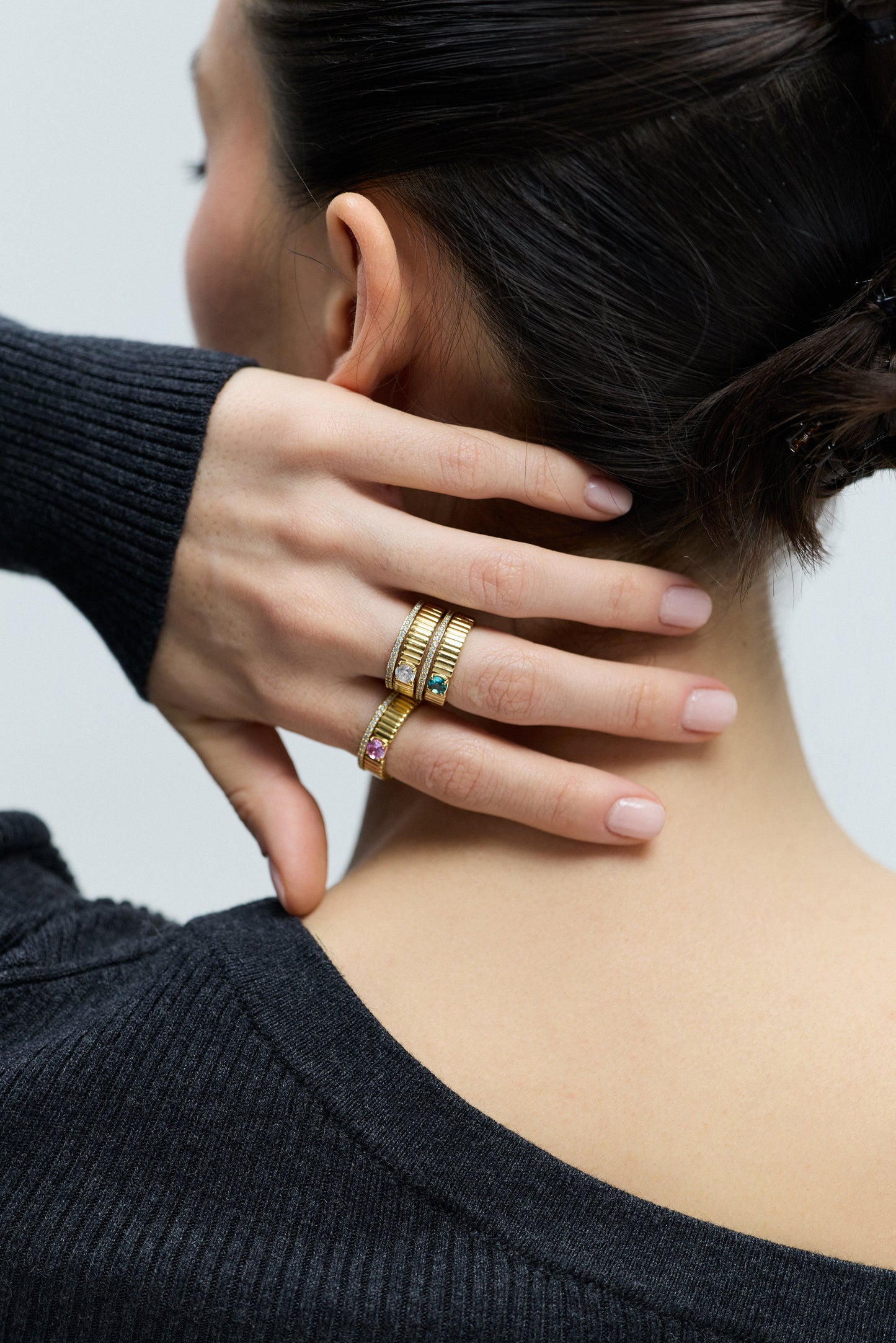 VIEW OF THE BACK OF A MODEL’S NECK WITH A TWISTED BRAID UPDO, SLIGHTLY TURNED TO THE SIDE WITH HER HAND RESTING AT THE BACK OF HER NECK, WEARING DELANCEY SOLITAIRE RING SETS ON HER FINGERS