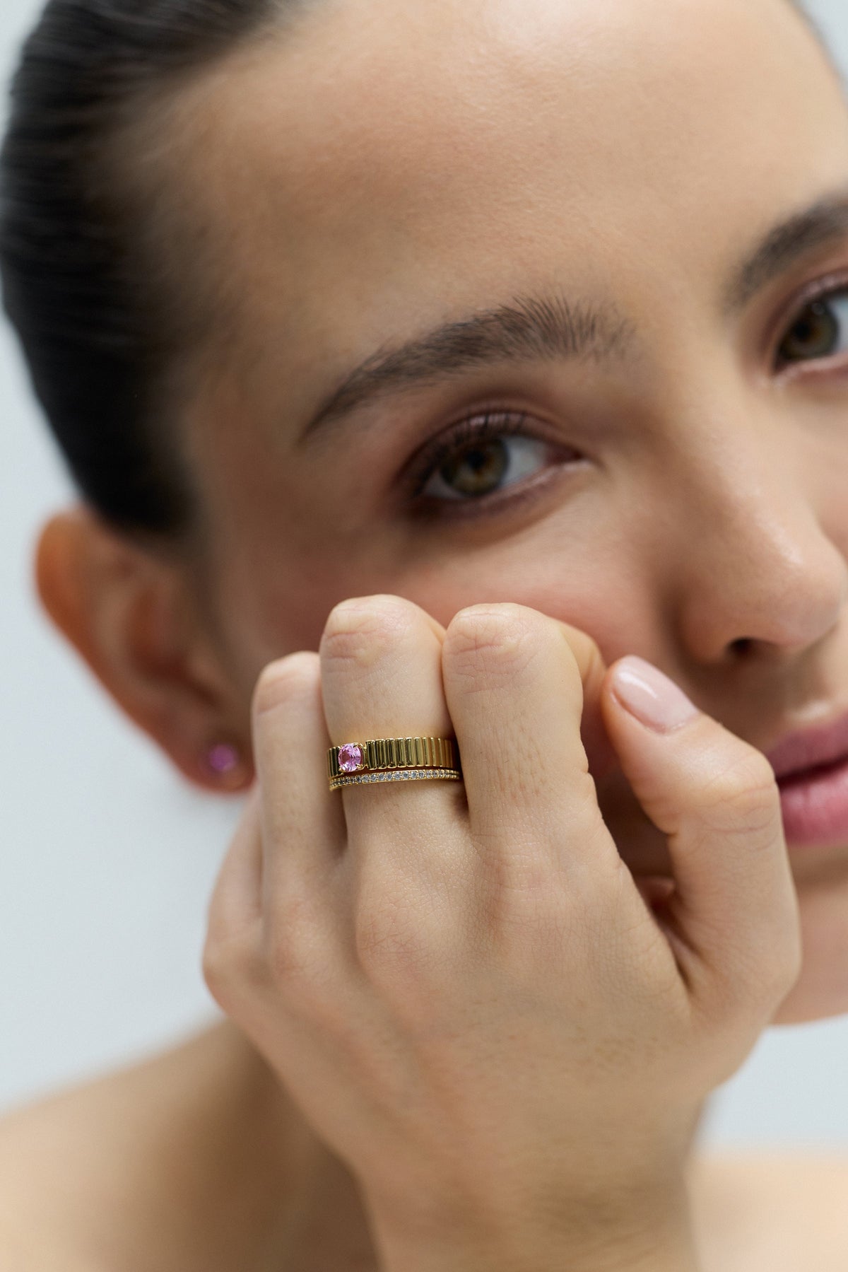 CLOSE UP OF A MODEL LOOKING AT THE CAMERA, RESTING HER CHIN ON HER CURLED HAND, WITH HER HAND IN FOCUS WEARING THE DELANCEY PINK SOLITAIRE RING