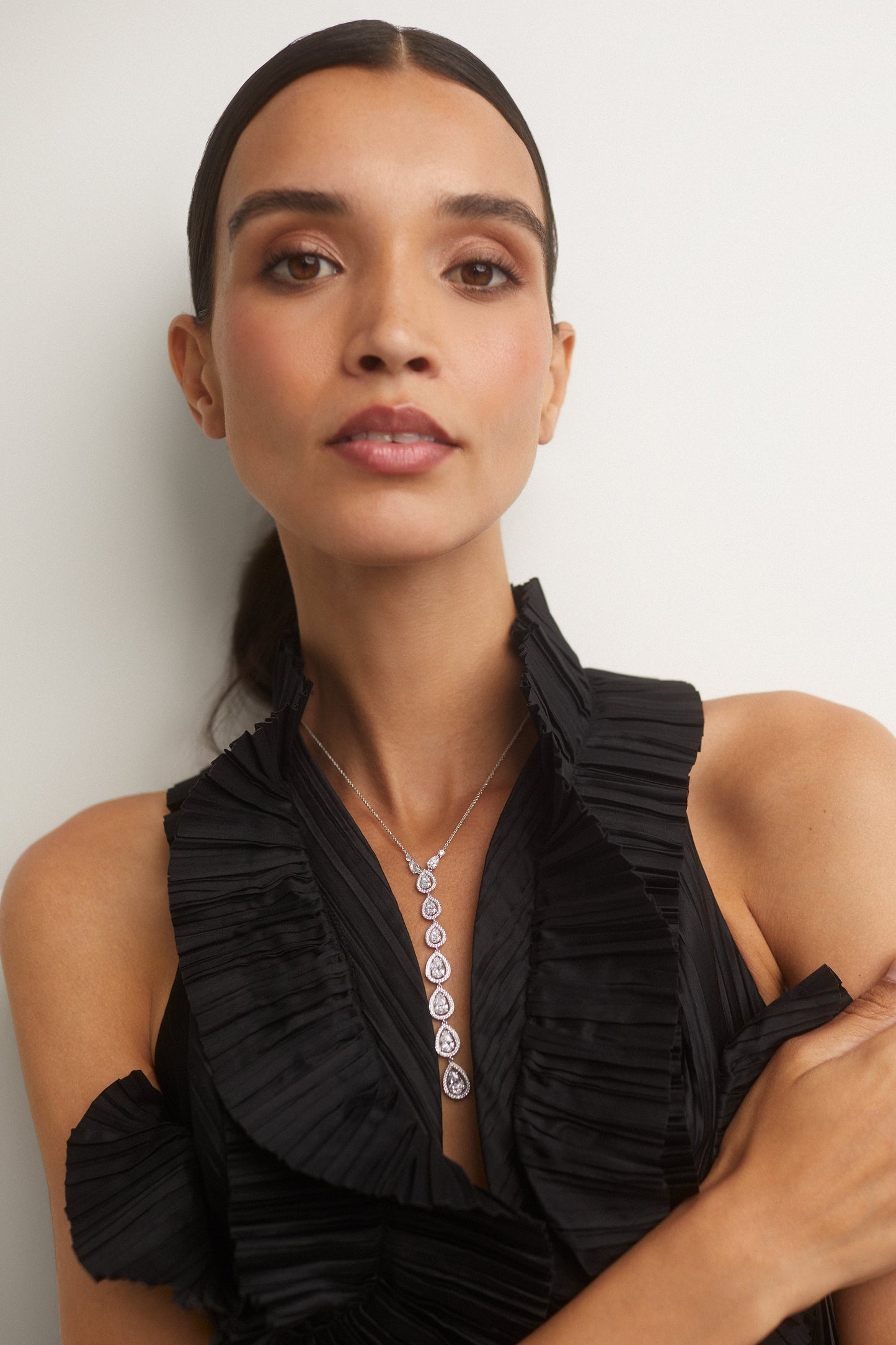 IMAGE OF A MODEL WEARING OUR RADIANT LONG PEAR CZ Y NECKLACE WITH A RUFFLY BLACK SLEEVELESS TOP.  SHE IS LOOKING DIRECTLY INTO THE CAMERA WITH HER ARMS CROSSED AND HER DARK HAIR IS PULLED BACK IN A SLEEK PONYTAIL.